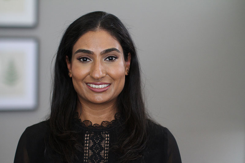 Dr. Sheikh of Romeoville Dental Center with long, dark hair wearing a black top with a lace pattern, standing against a plain wall with two framed pictures.