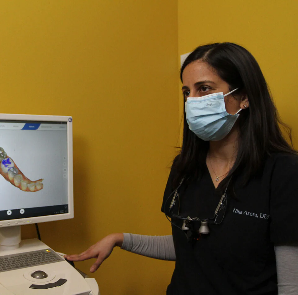 Dr. Nina Arora wears a face mask and reviews a dental scan on a computer screen, demonstrating patient care in a modern clinical setting.