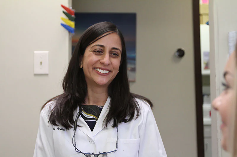 Dr. Nina Arora smiles while wearing a white coat and speaking with a patient inside Romeoville Dental Center, creating a warm and welcoming clinical setting.